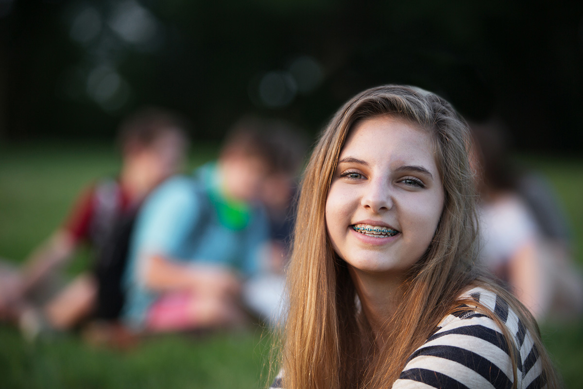 Teen girl smiling with braces in front of friends sitting on the grass.