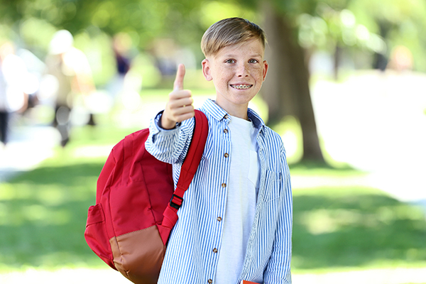 A child wearing braces and a red backpack giving a thumbs up.